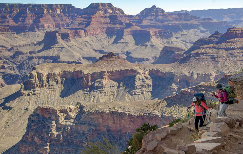 two backpackers with trekking poles are descending a unpaved trail into a desert landscape of colorful peaks, ridgelines and cliffs.