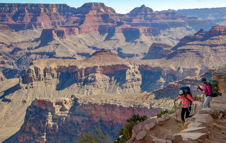 Whenever I hike the trails, even if I’m only intending to go a short distance, I always bring the ten essentials. At Grand Canyon plans can change quickly due to the weather, illness, injury or fatigue. two backpackers with trekking poles are descending a unpaved trail into a desert landscape of colorful peaks, ridgelines and cliffs.
