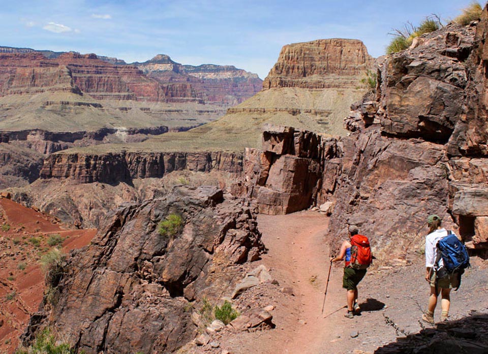 two backpackers are descending a unpaved trail in a desert landscape of colorful peaks, ridgelines and cliffs.