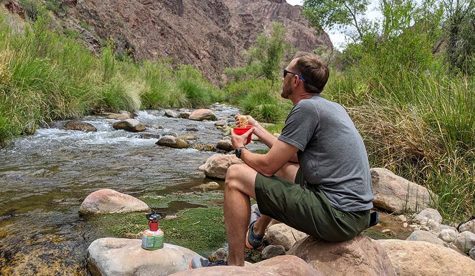 a person eating noodles out of a cup while sitting on a large slab of rock on the edge of a creek. In the distance, canyon cliffs.