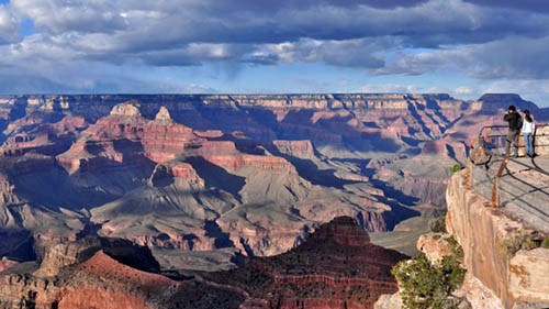 Two people behind a guardrail on the right looking at colorful peaks within Grand Canyon