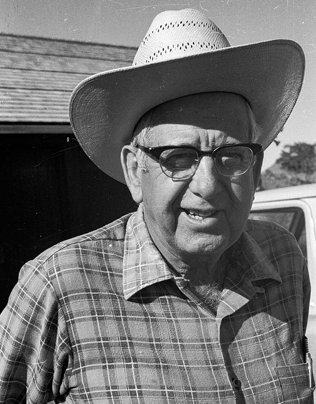 PORTRAIT OF LONG-TERM TUWEEP RANGER, JOHN RIFFEY, WEARING A STRAW COWBOY HAT, GLASSES AND A CHECKERED SHIRT.