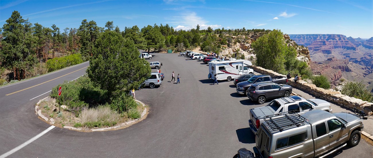 Alongside a two-lane road is a paved parking area with several cars and trucks parked next to a scenic viewpoint. In the distance, a colorful landscape of peaks and cliffs.
