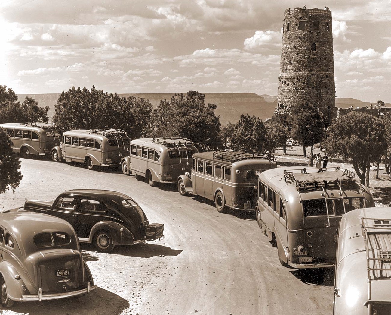 A sepia toned historic photo of six 1930's tour buses parked around a circular driveway with a cylindrical stone tower, several stories high, in the background.