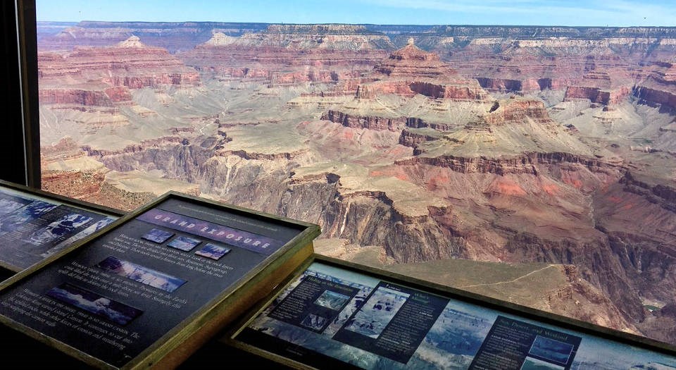 Looking past a row of geologic exhibits with text and photos, through a large glass picture window at a colorful Grand Canyon Landscape.