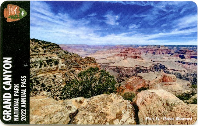 Text in black bar: Grand Canyon National Park 2022 Annual Pass. Photo shows a vast landscape with layers of red, green, and tan rocks, and a bright blue sky with wispy white clouds.