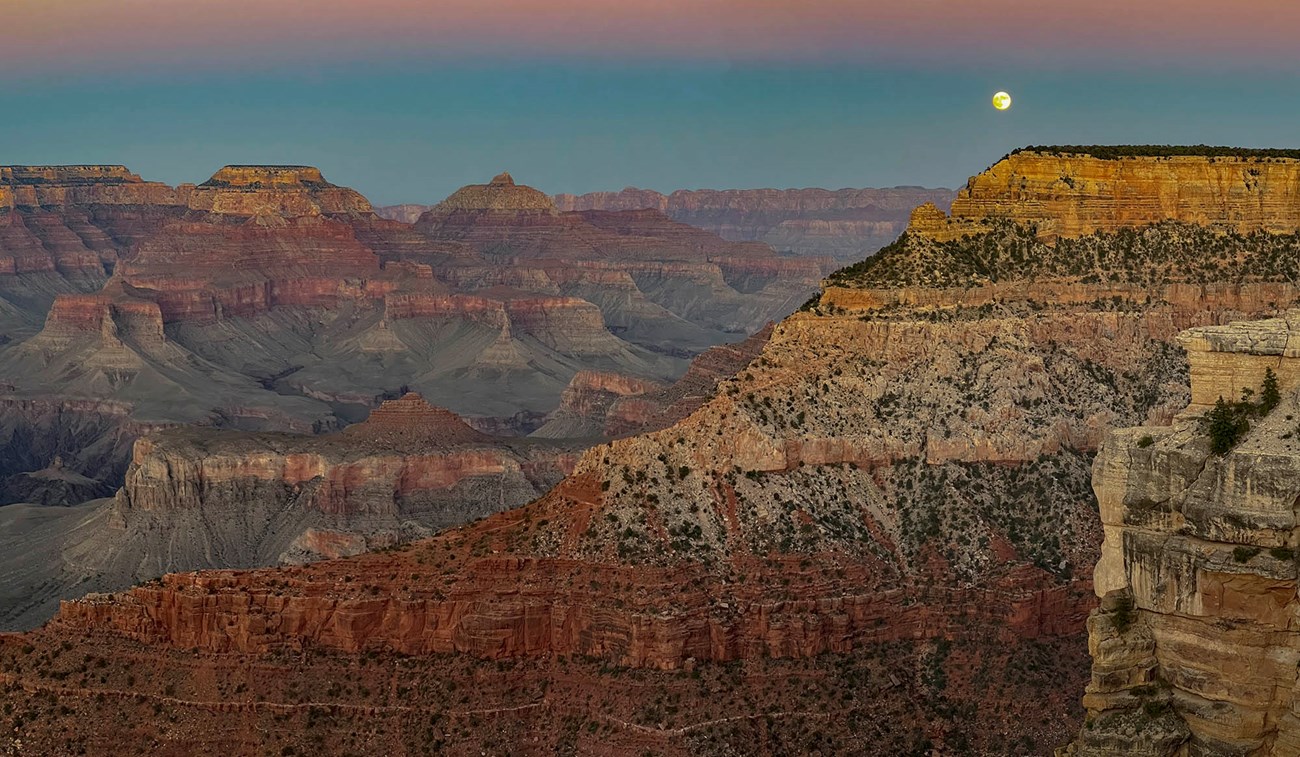 During twilight, a full moon is seen rising just above the horizon of a vast canyon landscape of colorful peaks and cliffs.