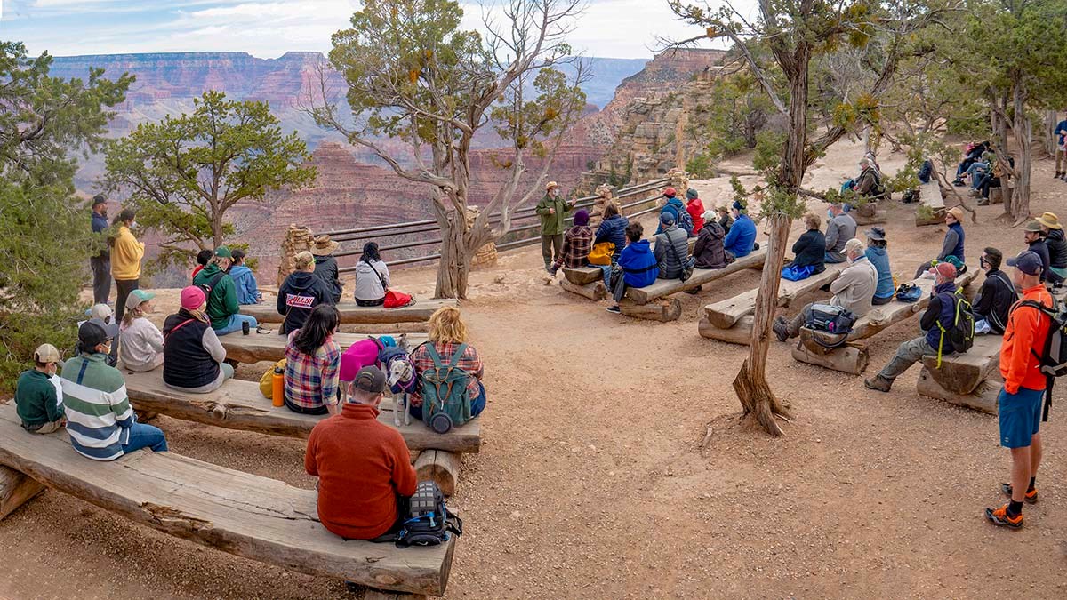a park ranger presenting a talk at an outdoor amphitheater. Visitors are sitting on wooden benches. canyon landscape in background.