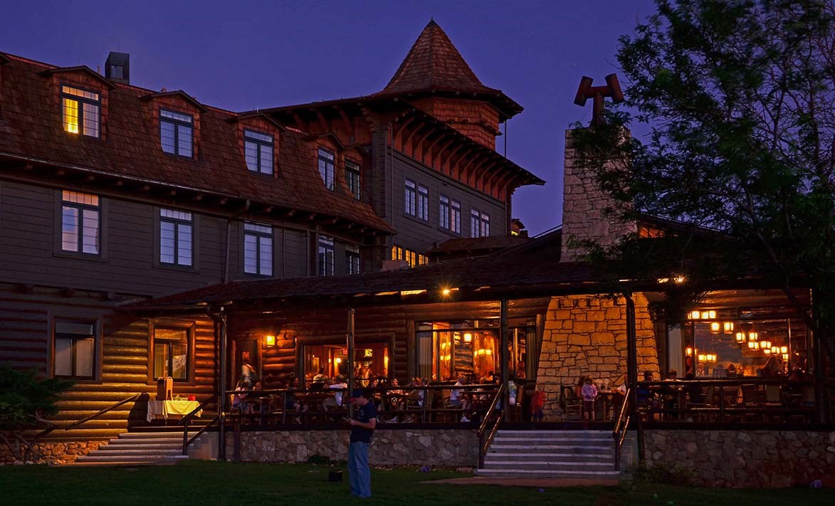 During twilight, a rustic stone and log lodge of three stories with people dining outside on a large covered porch that runs the length of the building.