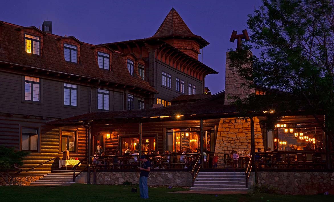 During twilight, a rustic stone and log lodge of three stories with people dining outside on a large covered porch that runs the length of the building.