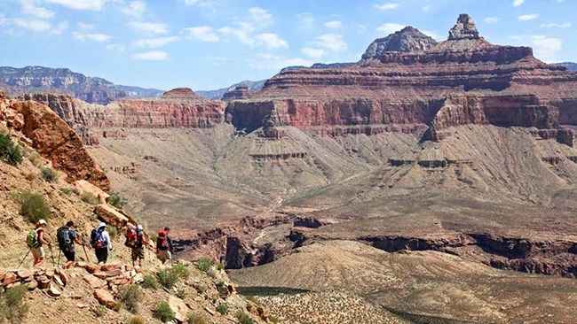 a group of 5 hikers walking down a backcountry trail into a desert landscape with a colorful peak formed from stratified rock layers on the right.