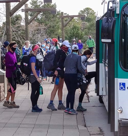 Just after sunrise, hikers with packs are boarding a white and green bus. Bus marquee reads, Hiker's Express.