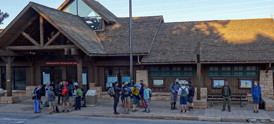 about 20 people with packs waiting at a bus stop in front of a rustic building with a gabled roof.