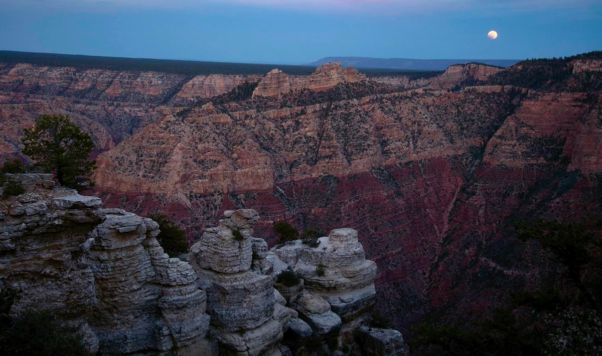 During twilight, eroded limestone pillars are in the foreground of a vast canyon landscape formed from layers of colorful rocks. A partially eclipsed full moon is rising above the horizon.