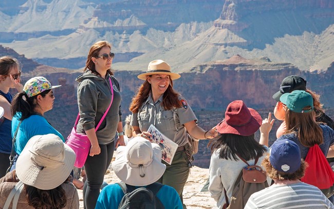 a smiling park ranger is talking to a group of visitors with the Grand Canyon landscape in the background.