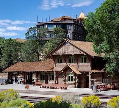 a historic log railroad depot with the words Grand Canyon above the main entrance. In the background, an historic hotel, several stories tall.