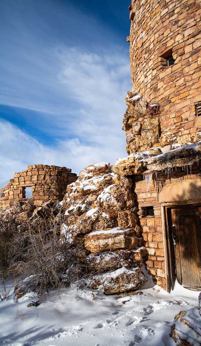 a portion of a round stone tower of rough-hewn masonry with snow covering the ground and a bright blue sky.
