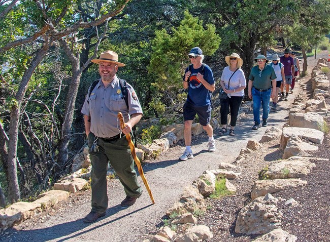 a park ranger with a large walking stick is leading a group of visitors down a paved footpath in a forested area.