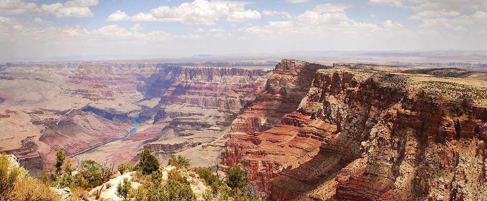 View north past tree-covered rocky outcrop towards green Colorado Rover in the distance. To the right are steep red-colored cliffs that extend from foreground, back along the river.