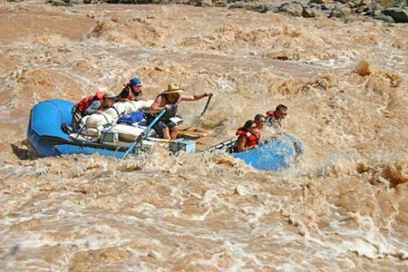 A boatman rowing a blue inflatable raft through a brown, churning Colorado River rapid. Four passengers are in the boat, two in front, and two in back.
