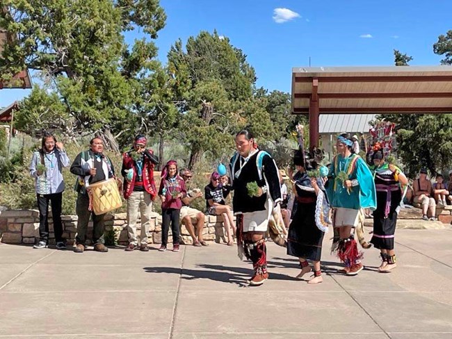 in an open air, paved plaza. four Hopi youth are dancing. In the background another group of four is chanting and drumming.