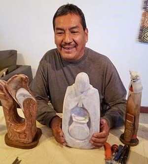 a middle aged Hopi Indian man sitting at a work bench and holding a stone sculpture of a human figure in front of him with both hands