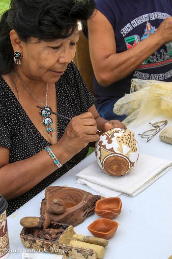 a woman sitting at a table and painting designs on a white vase. A display of small clay containers is in the foreground.