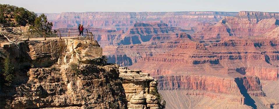 Two people standing behind the railings of a scenic overlook, with a vast canyon landscape of colorful cliffs and peaks in the distance.