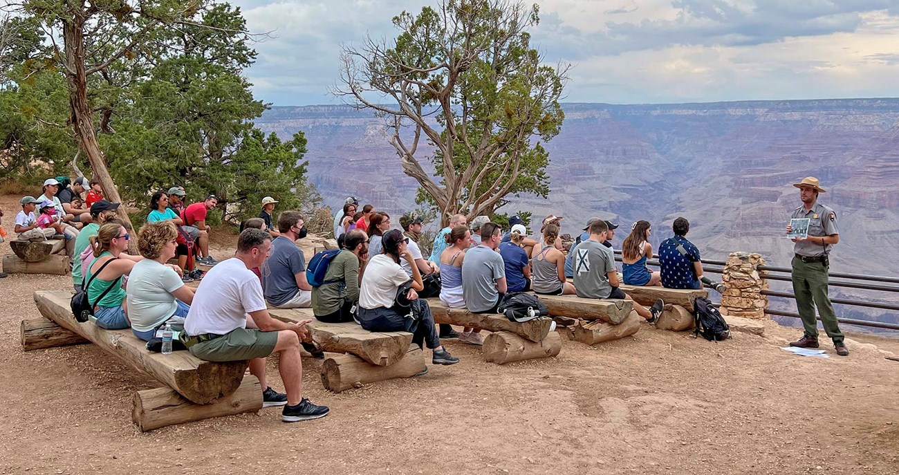 a group of around 40 park visitors wearing summer clothes and sitting on log benches are listening to a park ranger give a talk. He is standing on the far right and Grand Canyon is the background.
