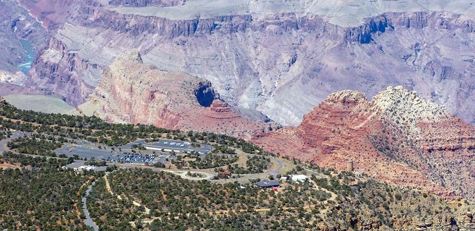 Aerial view of a forested plateau with a large parking area adjacent to a scenic overlook. In the distance, colorful vermilion peaks rise above a river within a rocky gorge.