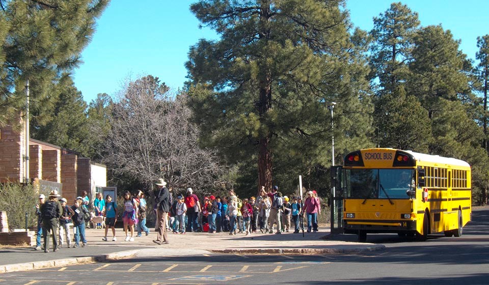 A yellow school bus parked alongside a curb in front of a building. Dozens of children and adults group outside.