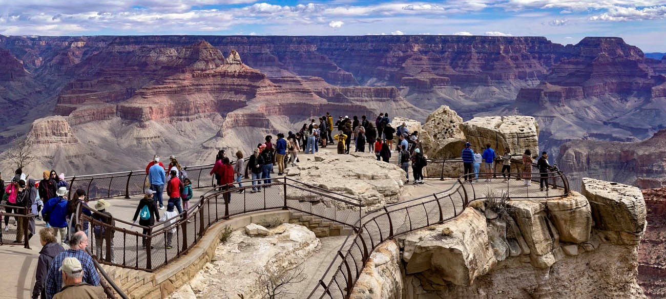 Midday view from a scenic overlook with a crowd of people; most are wearing jackets. They are interacting, and pausing to view a vast canyon landscape of colorful peaks and cliffs.