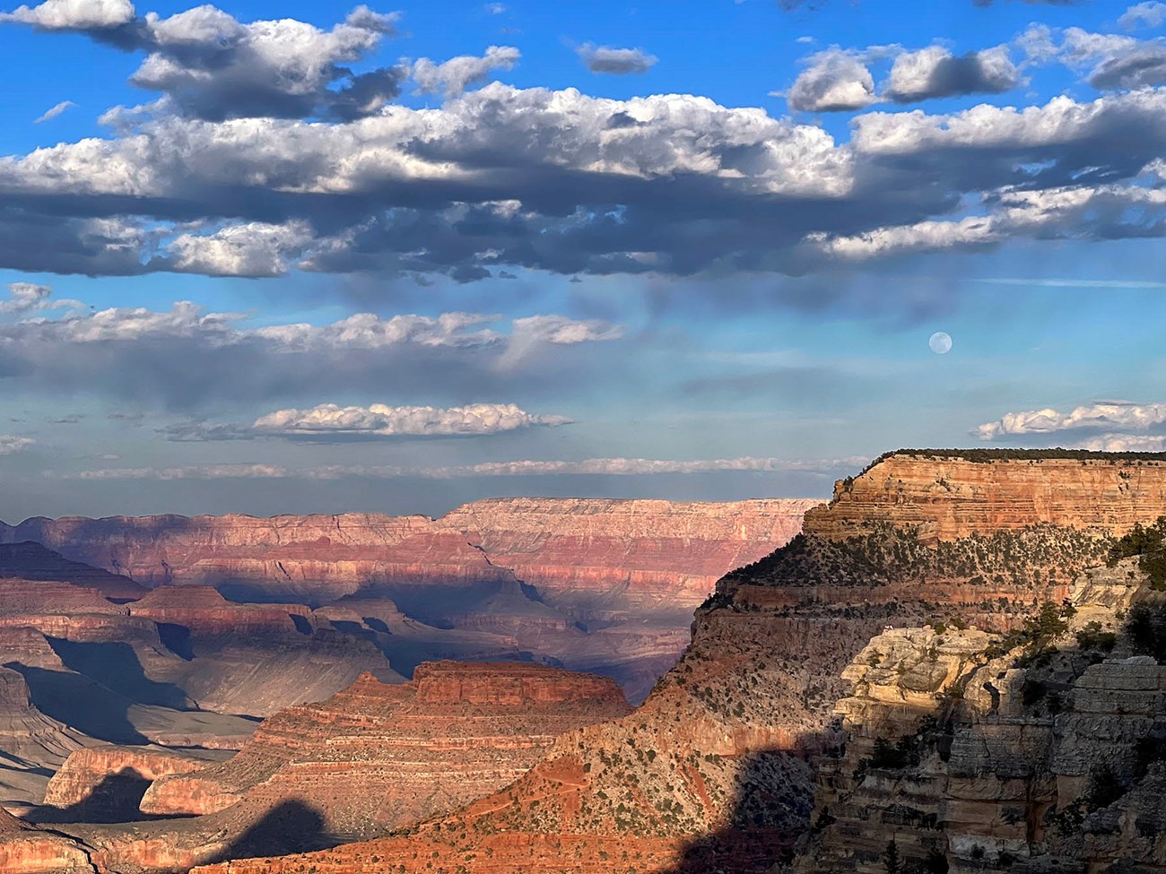 Approaching sunset, with lengthening shadows within a vast mile-deep canyon landscape. In a bright blue sky overhead, a full moon is rising between layers of cloud masses with dark undersides.
