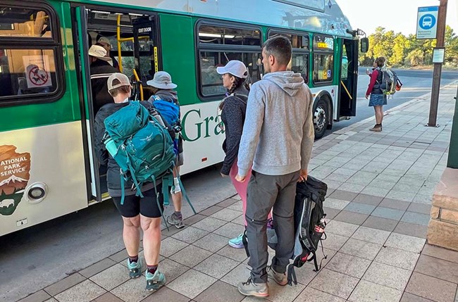 4 hikers with packs are boarding a white and green bus through the rear door.