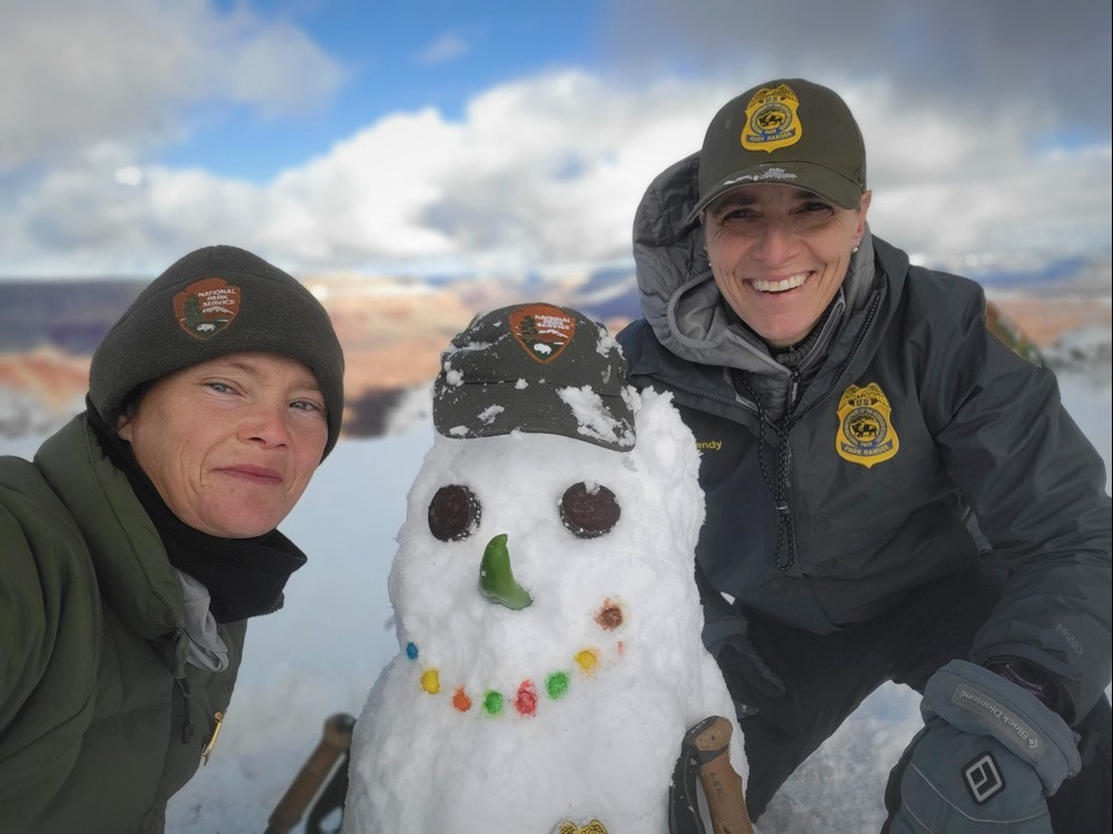 Two rangers pose with a created snow person on the rim of the canyon