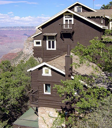 Looking past green oak trees at the west-facing side of Kolb Studio, several storied high, painted brown with white window frames. Grand Canyon is visible on the left and in the distance.