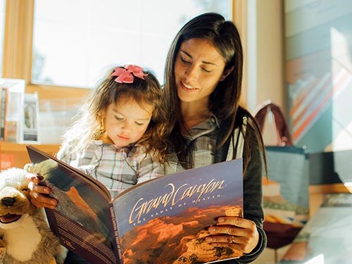 a mother is sitting next to her daughter, and they are looking at a large picture book entitled "Grand Canyon, the Vault of Heaven."
