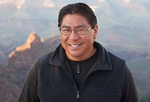 portrait of a middle aged Hopi Man with black hair and wearing a black jacket. Desert landscape in the background.