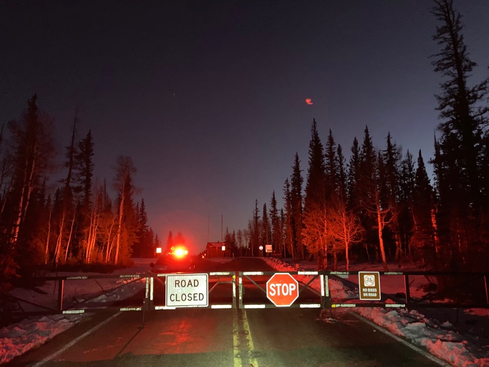 At dusk, a locked gate is closing off a two lane road at the park boundary - Signs, from left to right read; Road Closed, Stop, No Bikes On Road.