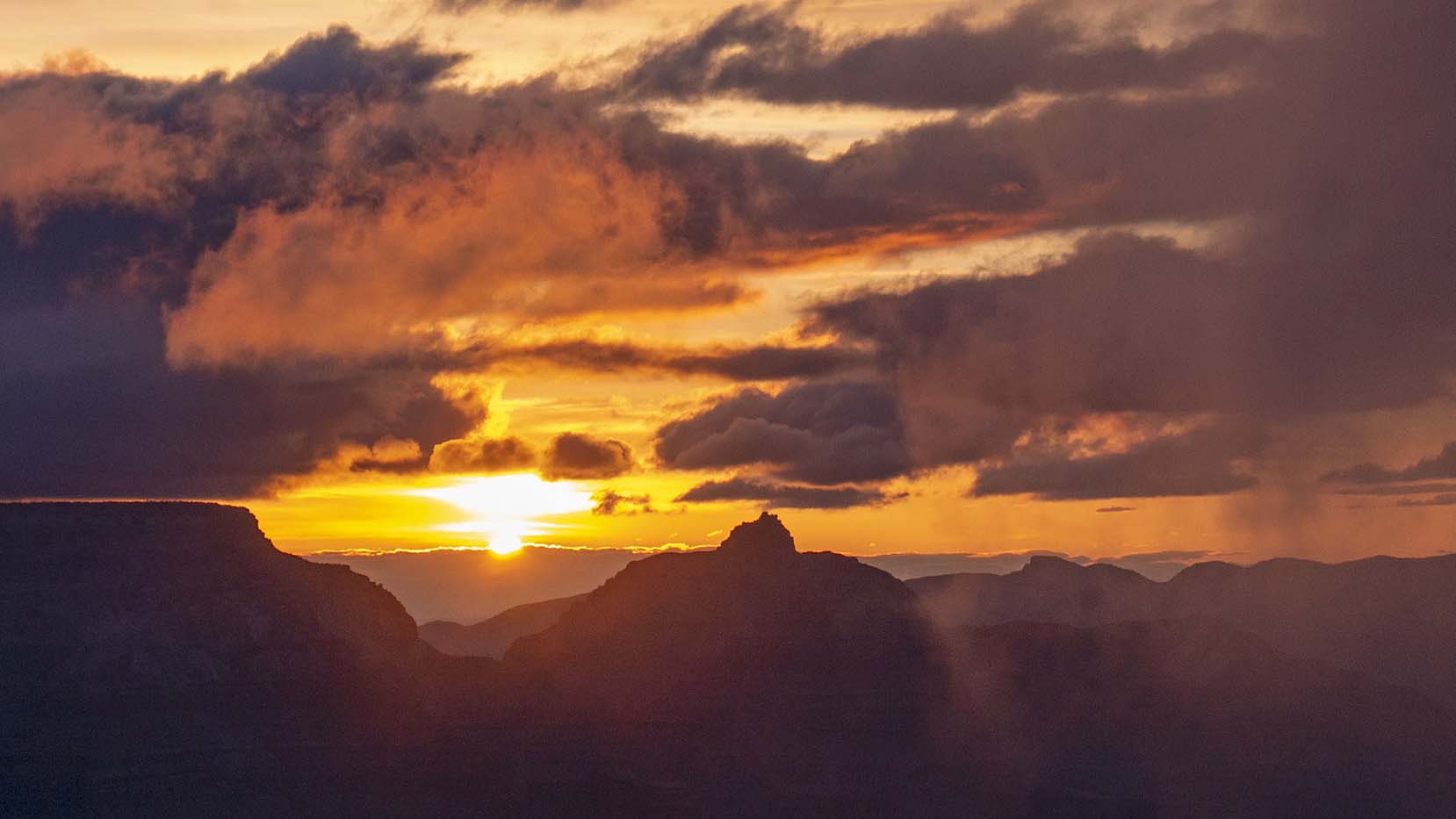 looking past silhouetted cliffs and peaks at the sun rising on the horizon, with purple and orange clouds filling the sky.