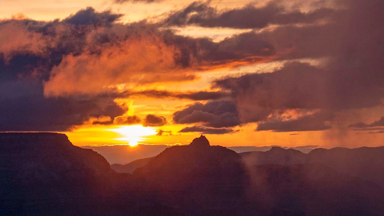 looking past silhouetted cliffs and peaks at the sun rising on the horizon, with purple and orange clouds filling the sky.