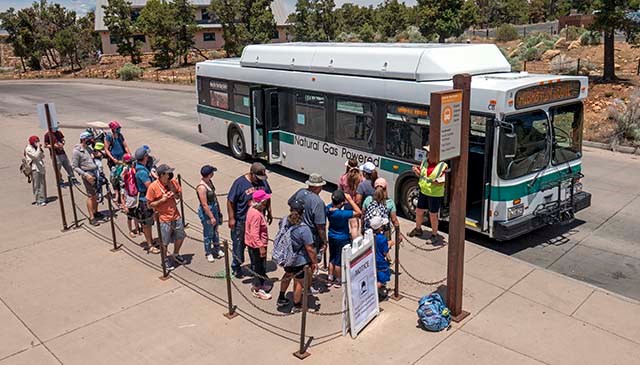 At a bus stop, a group of around 20 sightseers dressed in summer clothes are about to board a green and white bus.