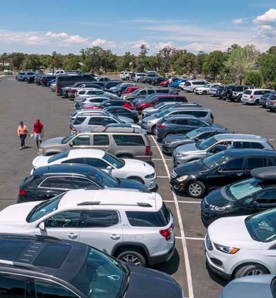 Several people walking past rows of cars in a busy parking lot with a pickup truck pulling out behind them