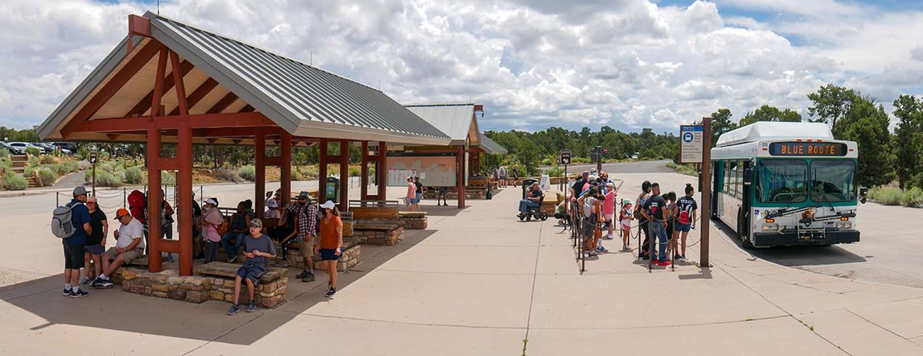 An outdoor shuttle bus terminal. In the foreground, about a dozen people are sitting under an open air shade pavilion. On their right, a line of people about to board a green and white bus.