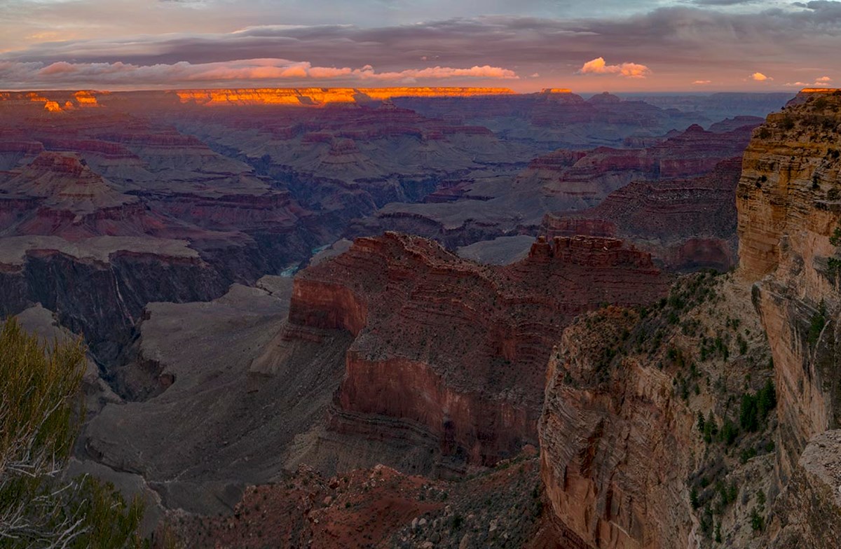 Looking down into a vast canyon landscape of colorful cliffs and peaks almost engulfed by twilight shadows. The distant horizon is illuminated by the final beam of sunset light. NPS/M. Quinn