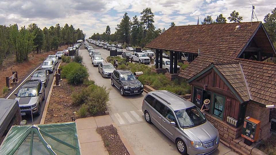 Four lines of cars wait in line to pay the entrance fee at a wooden building with gabled roofs.