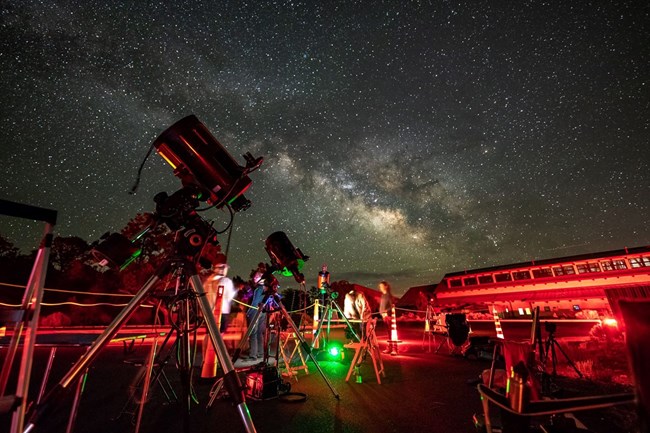 A telescope in the foreground aims up at a Milky Way spanning across the sky. Red lights decorate the ground below.