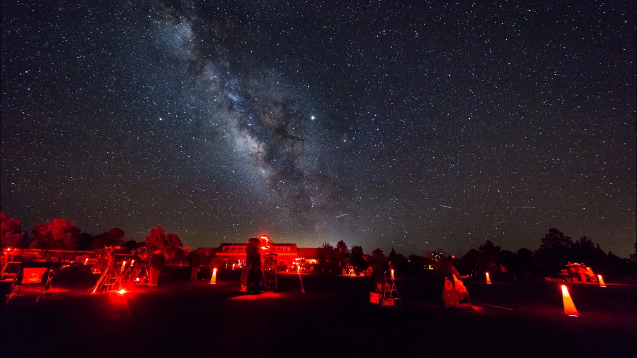 The white Milky Way over a red lit parking lot with people and telescopes.