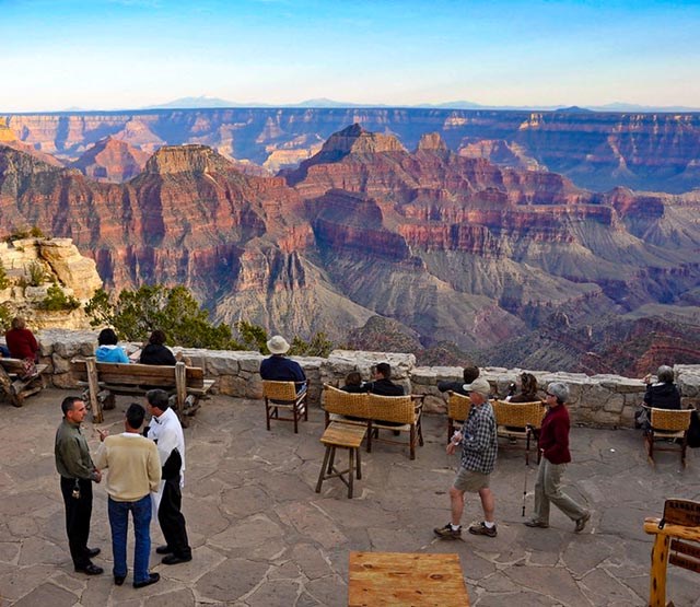 about a dozen people sitting on the flagstone veranda of a lodge and looking out at a series of connected peaks within a vast canyon landscape