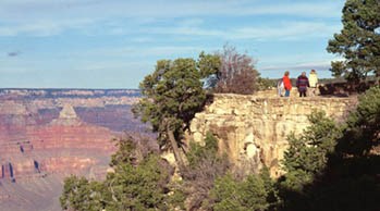 Several people are walking along the canyon rim behind a stone guard wall. The canyon is visible on the left Several people are walking along the canyon rim behind a stone guard wall. The canyon is visible on the left